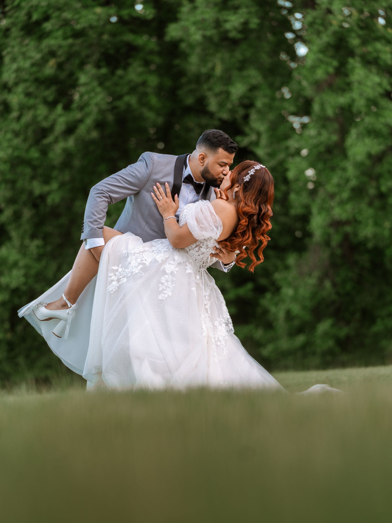 Groom in gray suit dips bride in white lace gown for a romantic kiss outdoors with lush green trees.