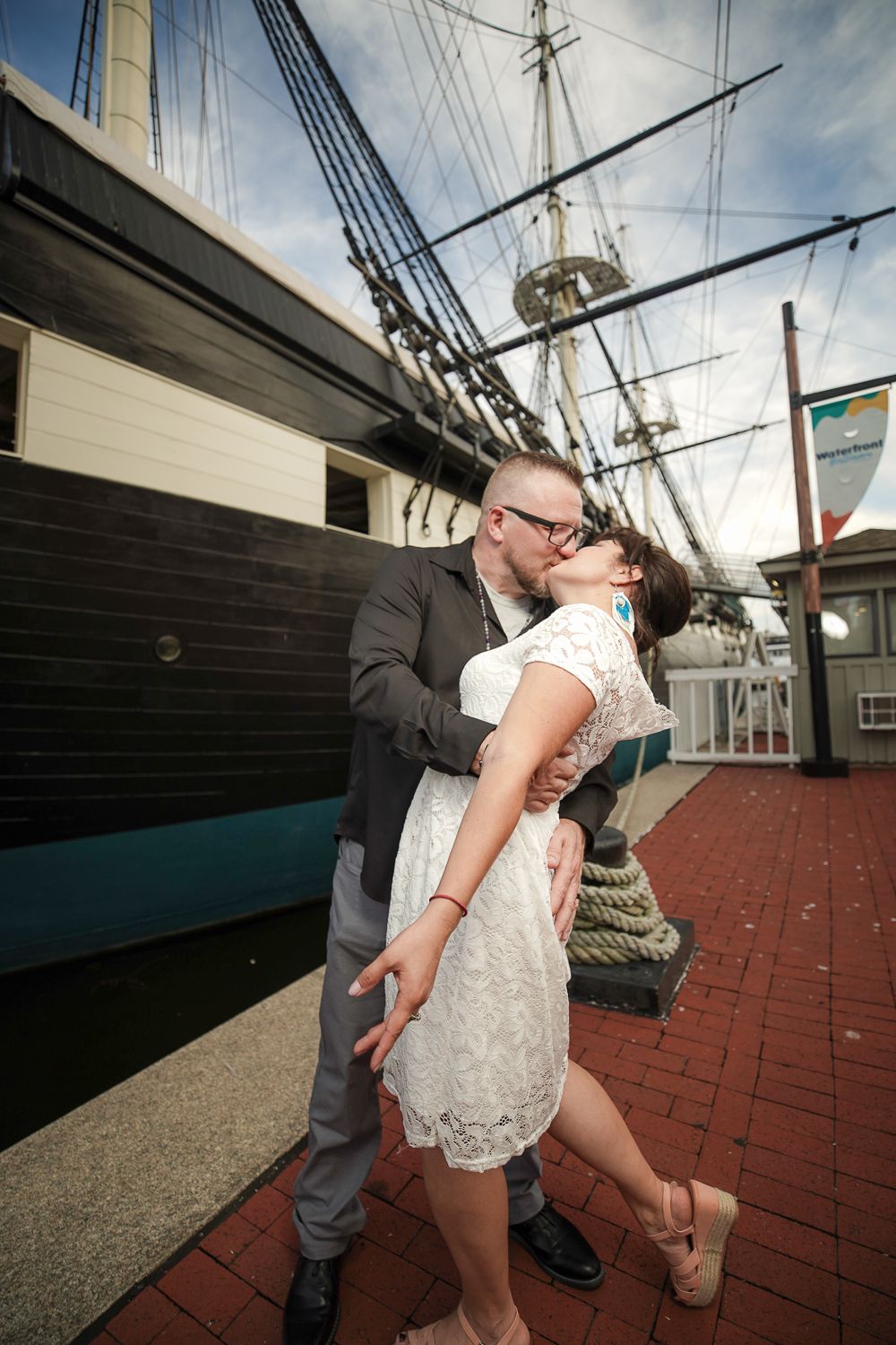 Newlywed couple sharing passionate kiss beside historic tall ship at Baltimore Inner Harbor.