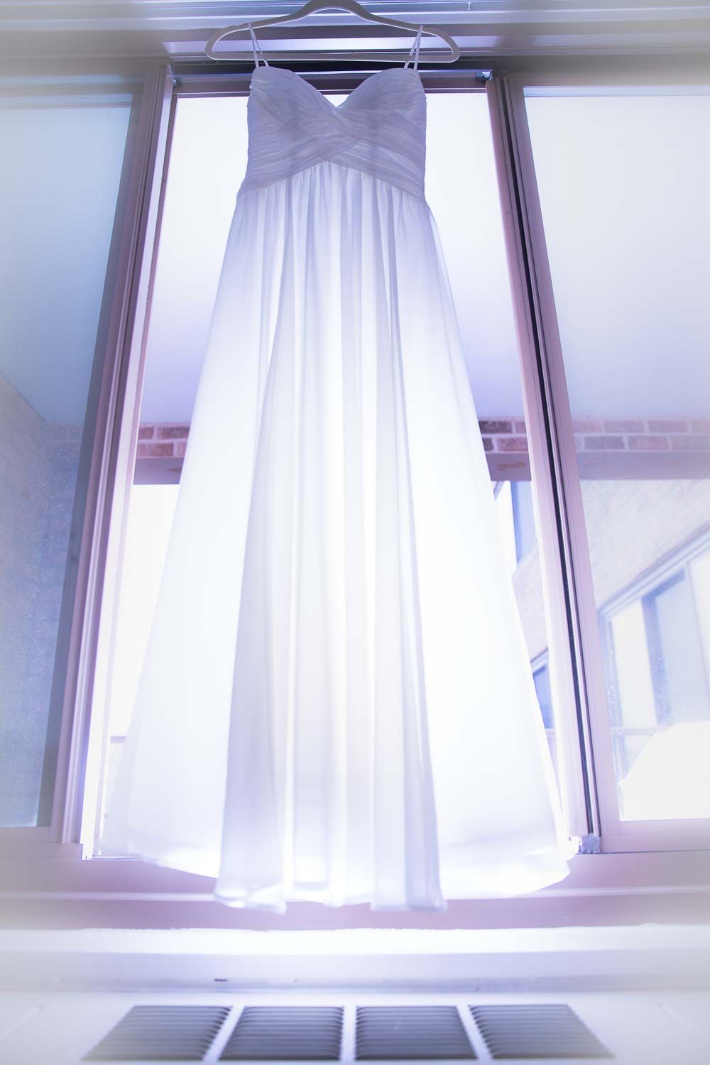White strapless wedding dress hanging on a hanger in front of a bright sunlit window.