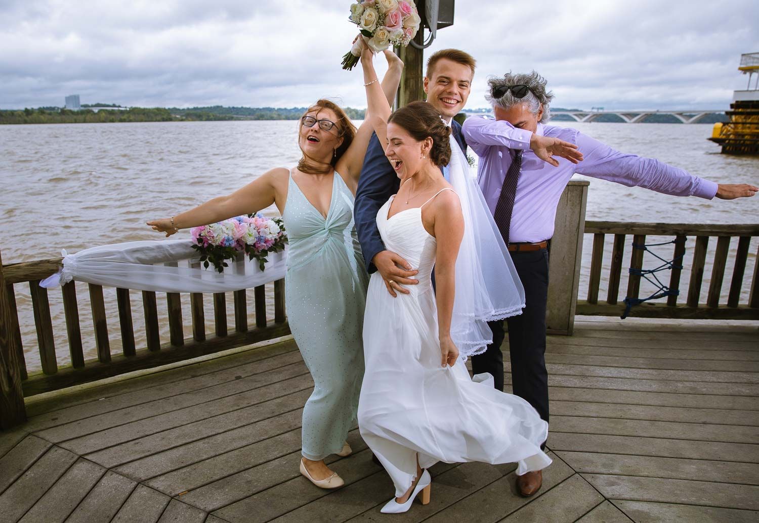 Joyful wedding party celebrating on a waterfront deck, bride tossing bouquet while guests cheer.