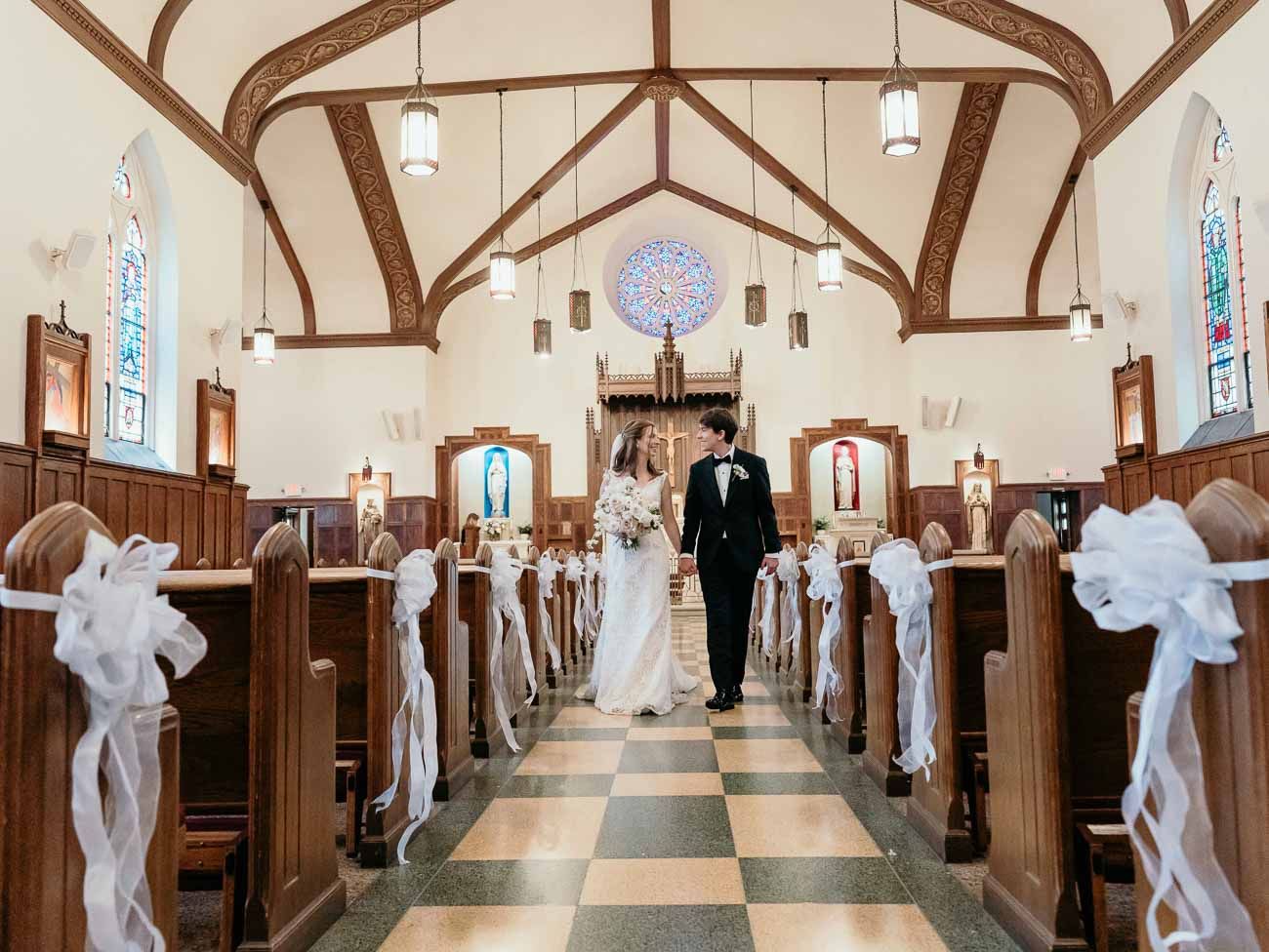Bride and groom walk together down a checkered aisle in a charming church with white bow decor.