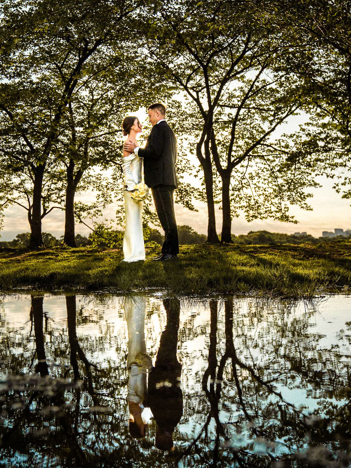 Bride and groom stand together near a reflective pond surrounded by trees in golden sunset light.