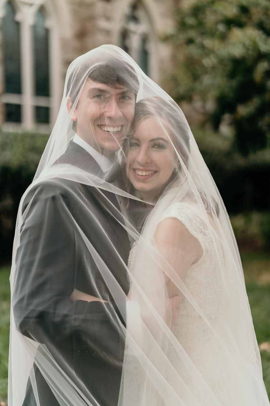 Smiling bride and groom pose together beneath the bride's sheer white veil outside a stone church.