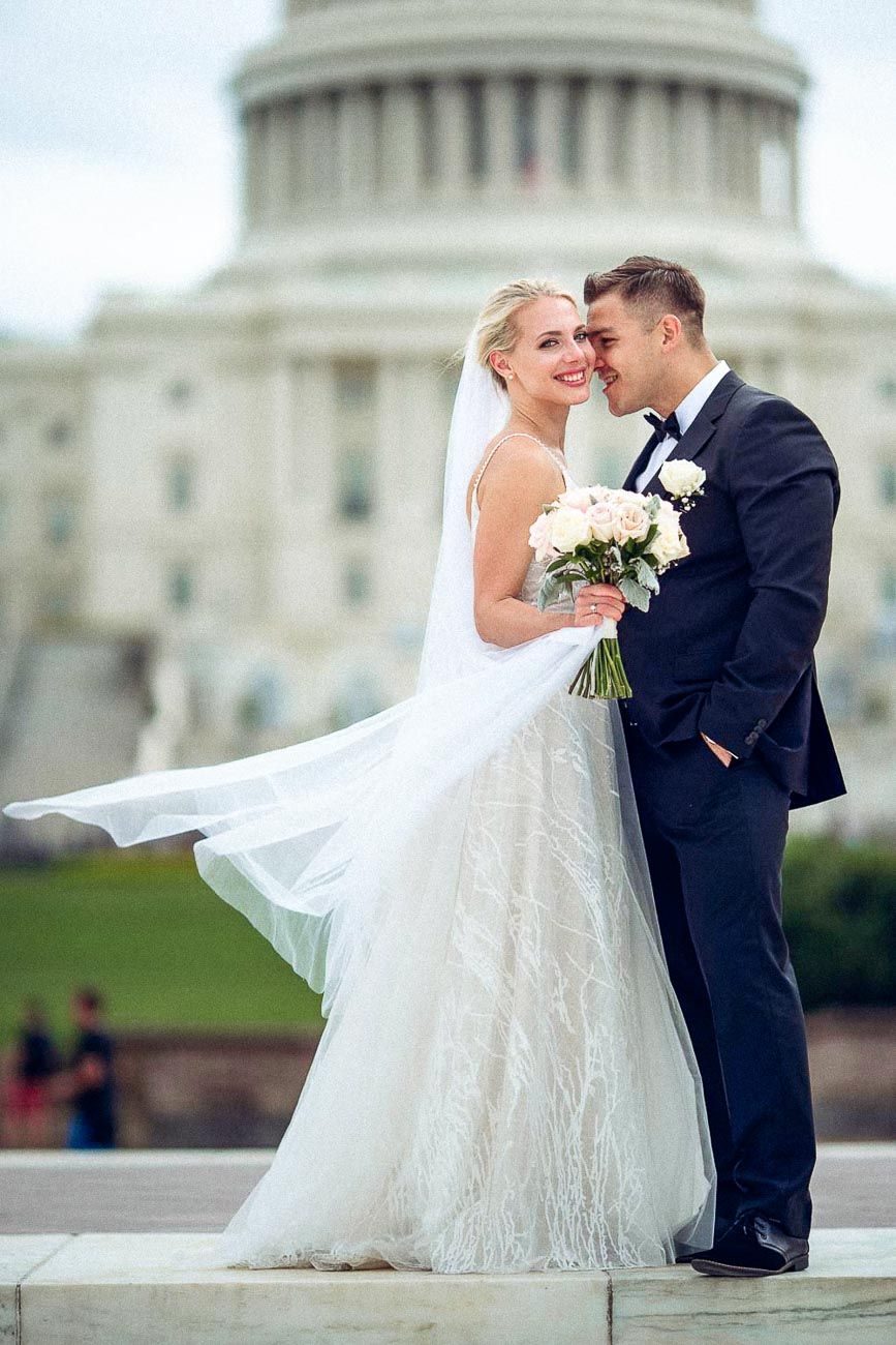 Bride and groom sharing a tender forehead touch in front of the US Capitol, bride's veil flowing in breeze.