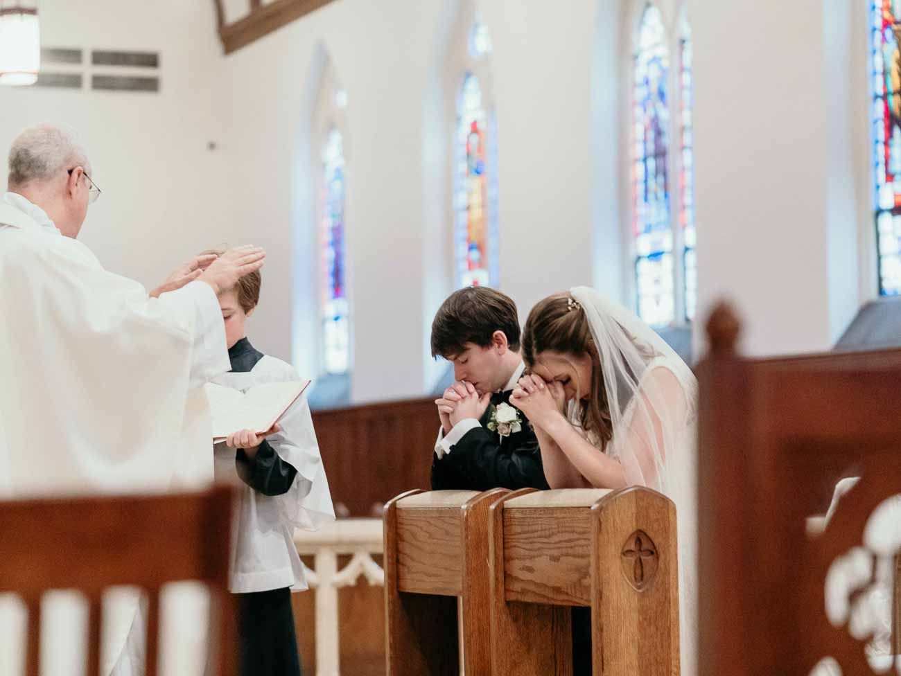 Priest blesses bride and groom as they bow their heads in prayer during a Catholic wedding ceremony.