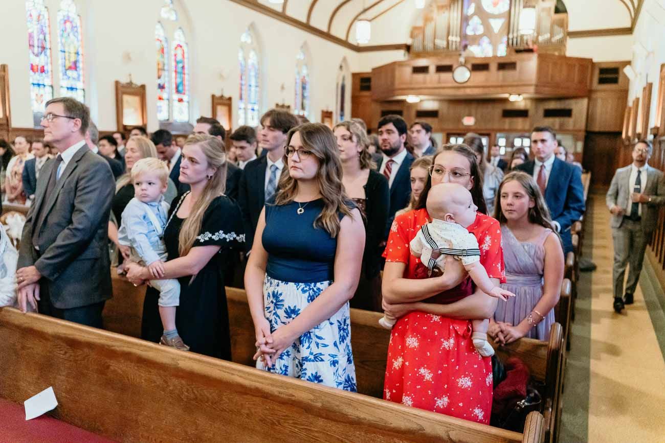 Wedding guests including families with babies sit and stand in wooden church pews during ceremony.