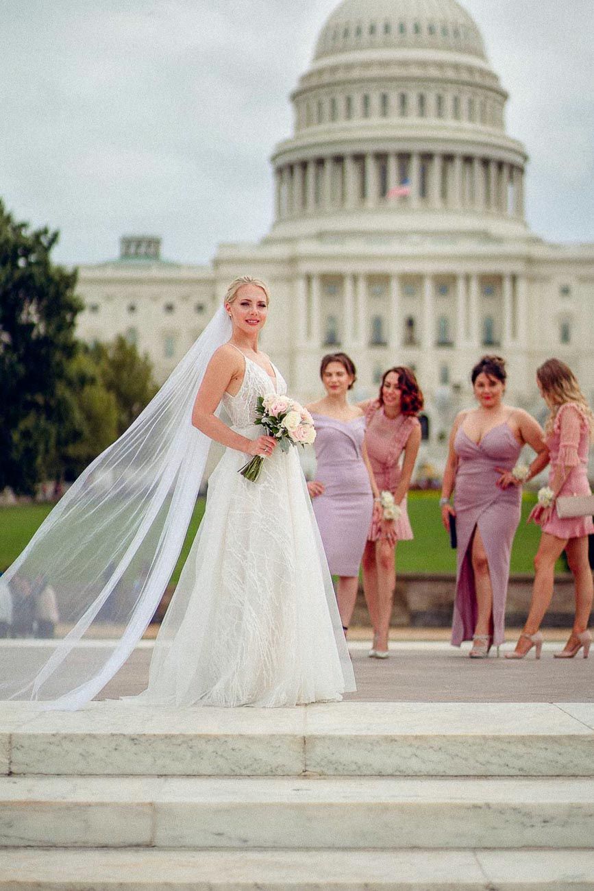 Elegant bride in white gown with long veil holding bouquet before the US Capitol with bridesmaids behind.