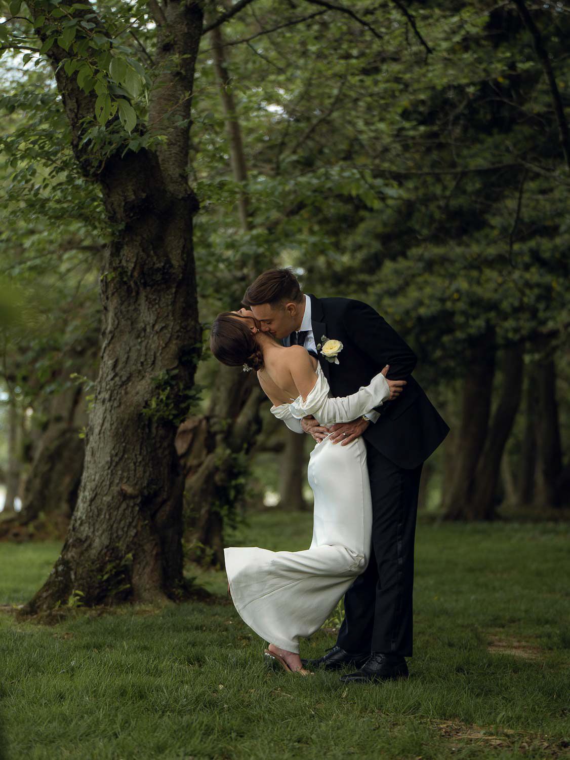 Groom dips bride for a kiss beside a tree in a lush green wooded park setting.