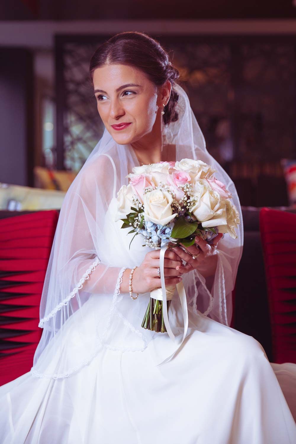 Beautiful bride in white gown and veil sitting while holding a romantic pink and white flower bouquet.