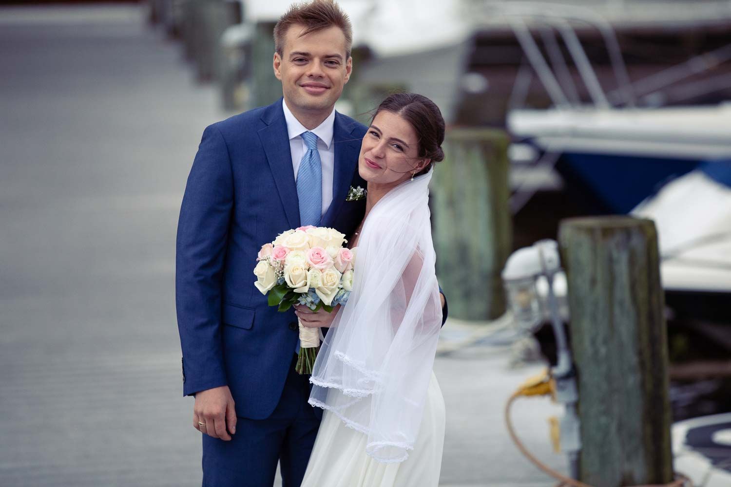 Smiling bride and groom posing at marina dock, bride holding pink bouquet in white gown.