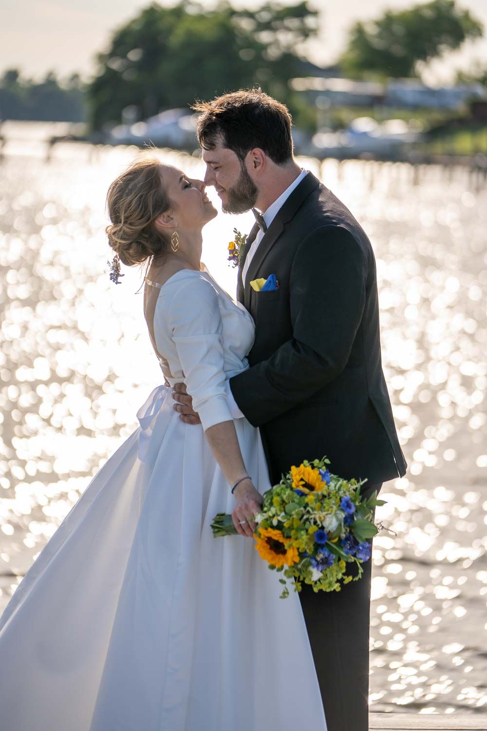 Bride and groom sharing romantic moment on waterfront dock, bride holding vibrant sunflower bouquet.