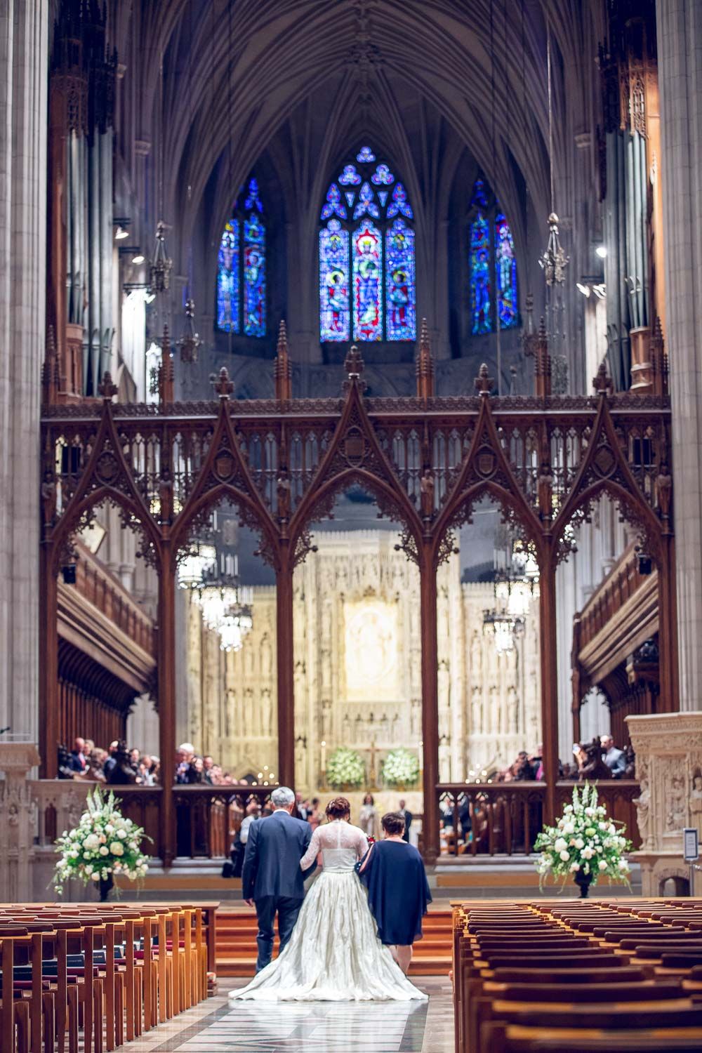 Bride walks down the aisle of a stunning Gothic cathedral with stained glass windows and floral arrangements.