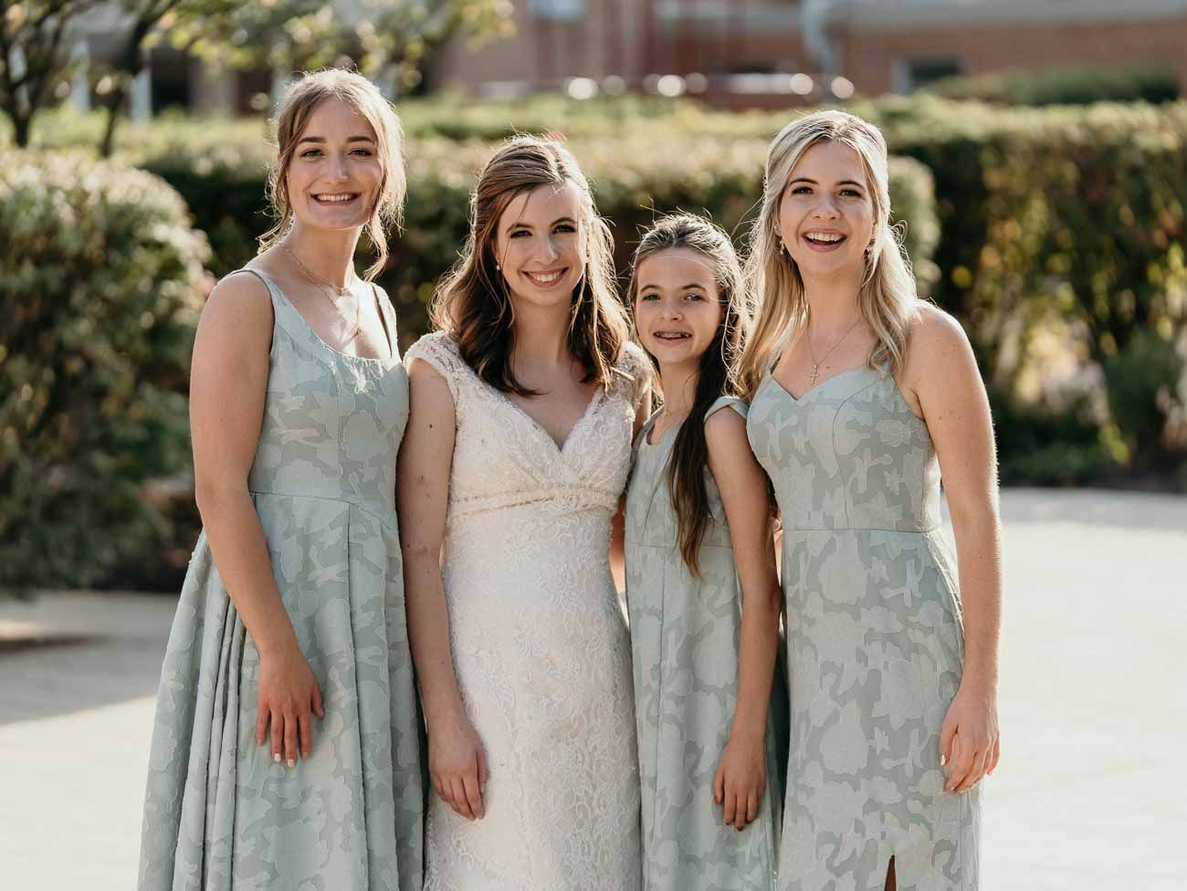 Bride in lace gown smiles with three bridesmaids in sage green floral dresses outdoors.
