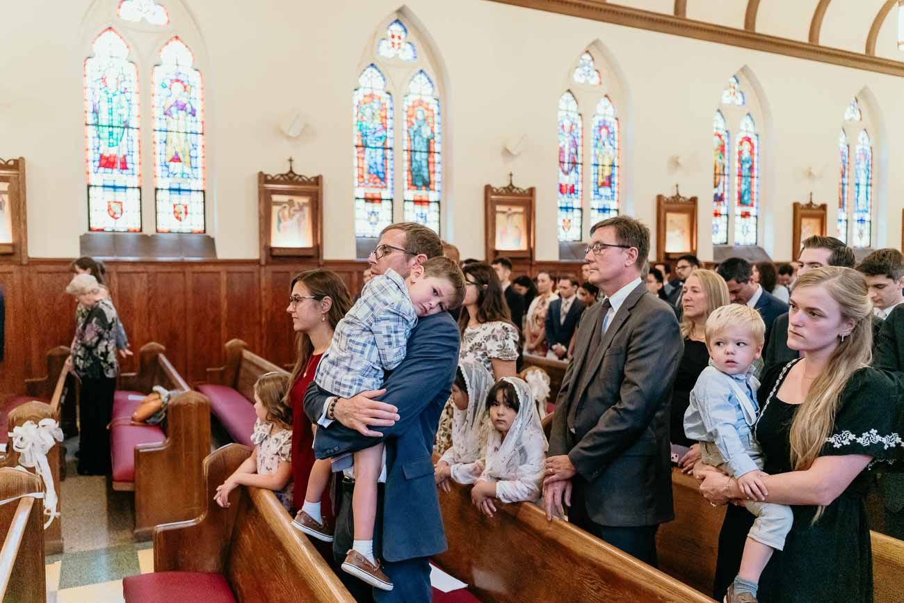 Wedding congregation with families and young children standing in pews of a stained glass church.
