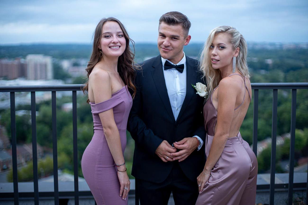 Groom in black tuxedo posing with two bridesmaids in mauve dresses on a rooftop with city views behind.