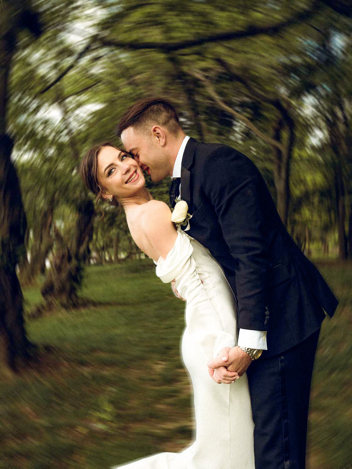 Groom dips bride in white gown while sharing a joyful moment in wooded area.