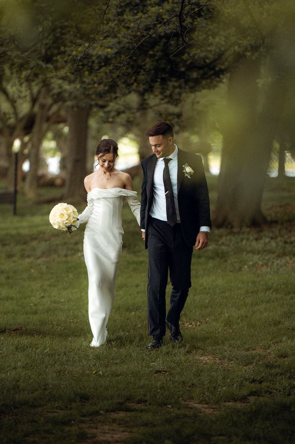 Bride and groom walk hand in hand through a tree-lined path in a lush green park.