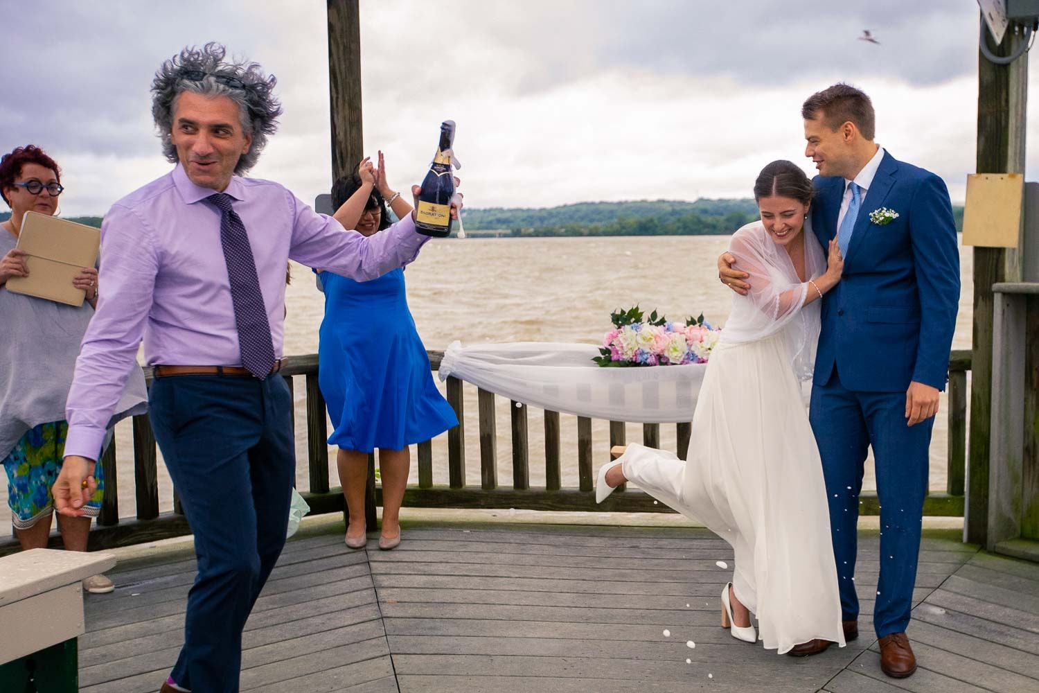 Joyful wedding celebration on a waterfront deck with guest holding champagne bottle near bride and groom.