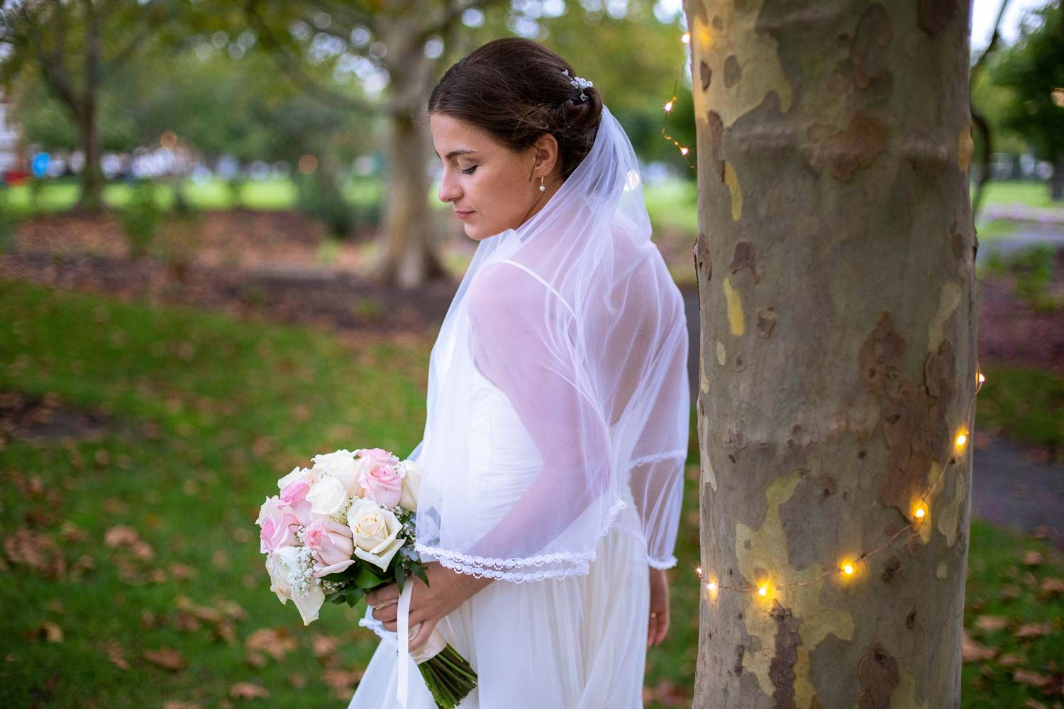 Bride in white gown and veil holding pink and white rose bouquet standing by a tree with fairy lights.