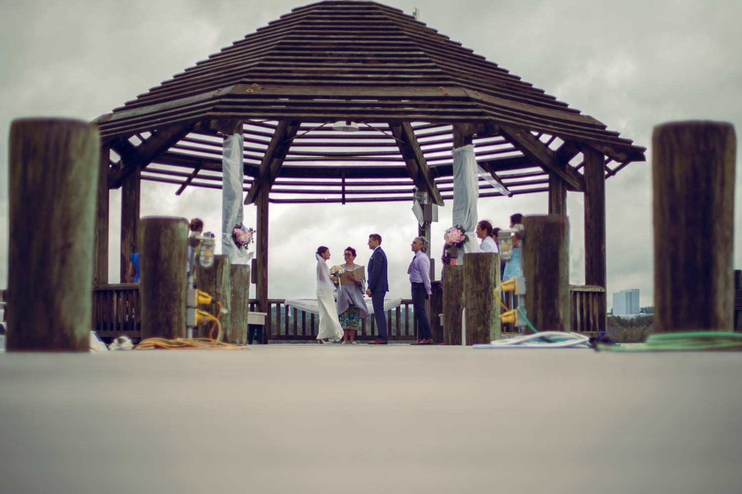 Distant view of small wedding ceremony gathering under a rustic wooden gazebo on a waterfront pier.