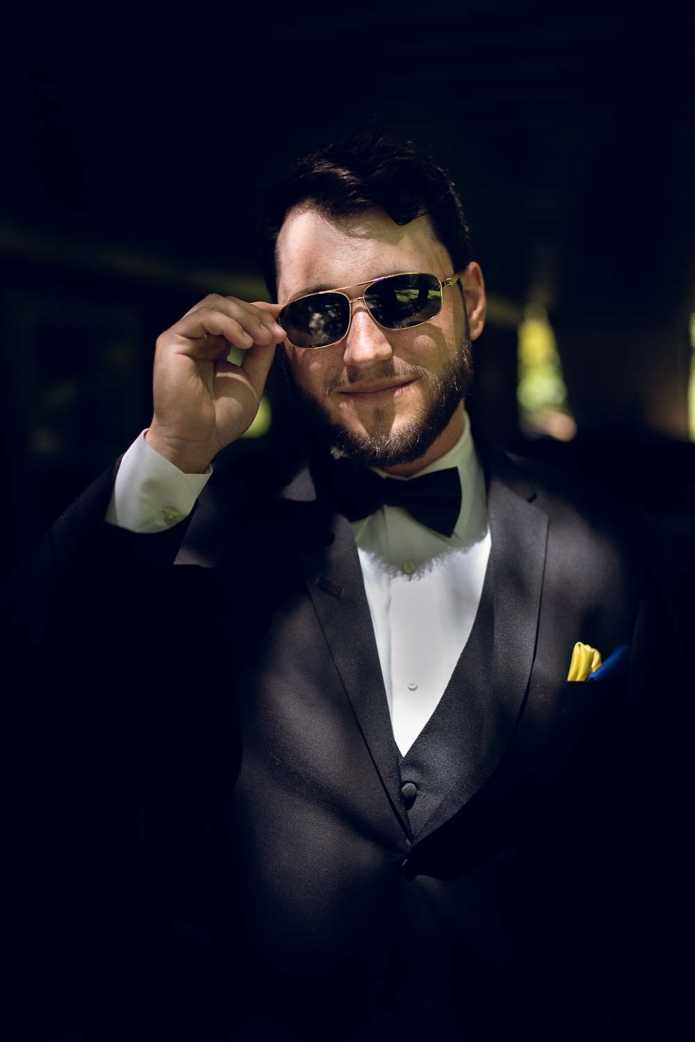Stylish groom in black tuxedo and bow tie smiling while lowering sunglasses at reception.