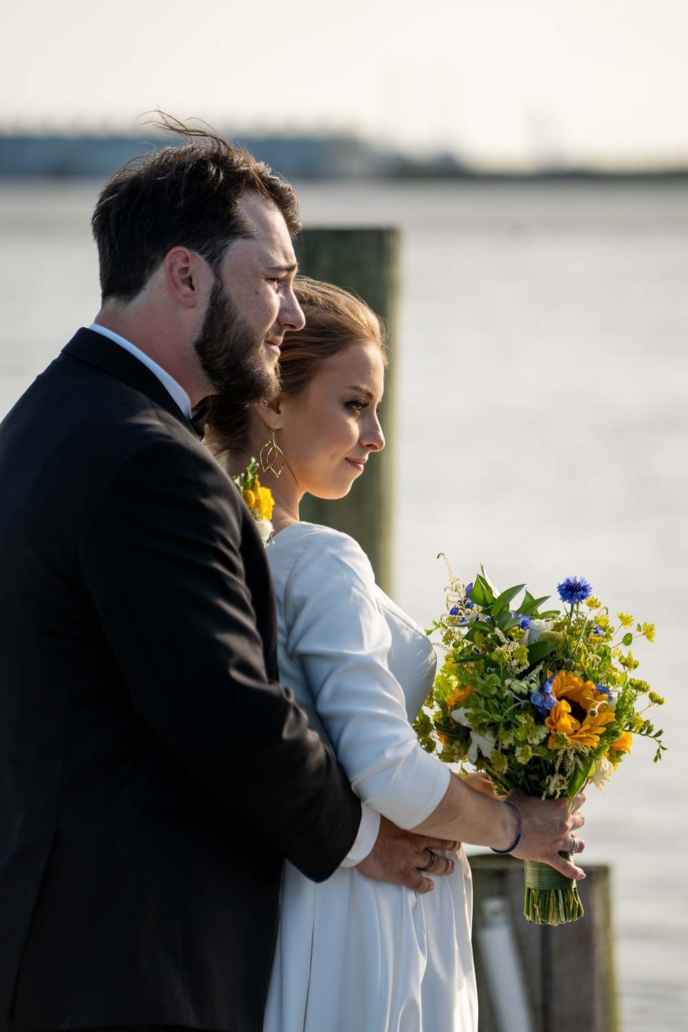 Bride and groom standing together on waterfront dock, holding colorful wildflower wedding bouquet.