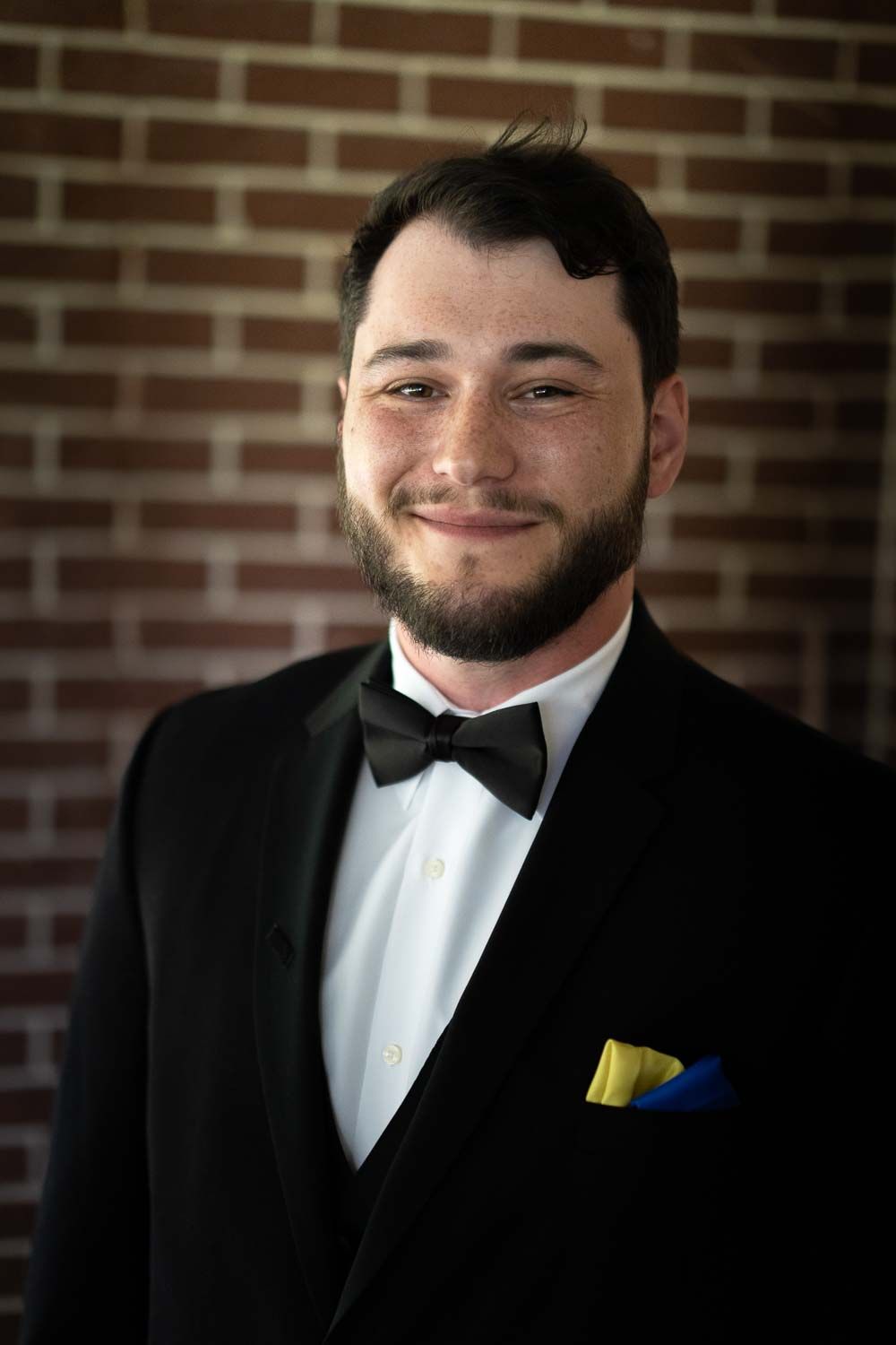 Smiling groom in a black tuxedo with bow tie and yellow pocket square against a brick wall.
