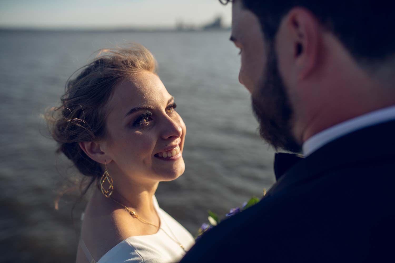 Radiant bride gazes lovingly at groom near sparkling water during golden hour wedding portraits.