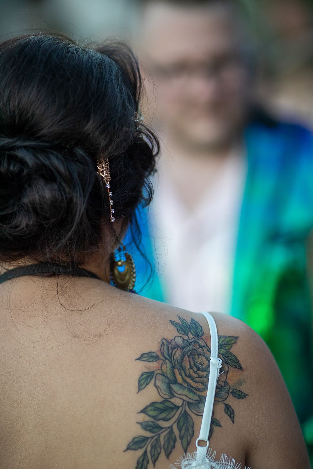 Close-up of bride's elegant updo and rose shoulder tattoo with man in green sequin jacket blurred in background.