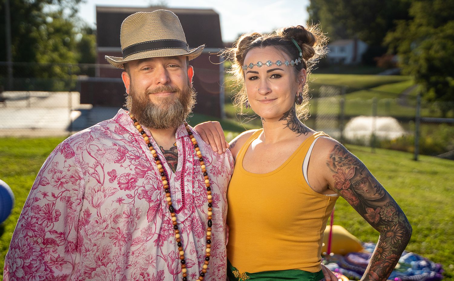 Couple standing together outdoors, man in floral hat and woman with tattoos.