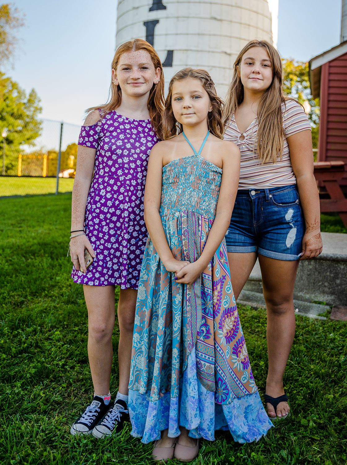 Three young girls posing outdoors in summer dresses near a silo on a sunny day.