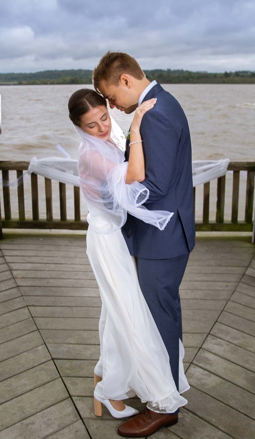 Bride in white gown and groom in blue suit share intimate embrace on waterfront wooden deck.