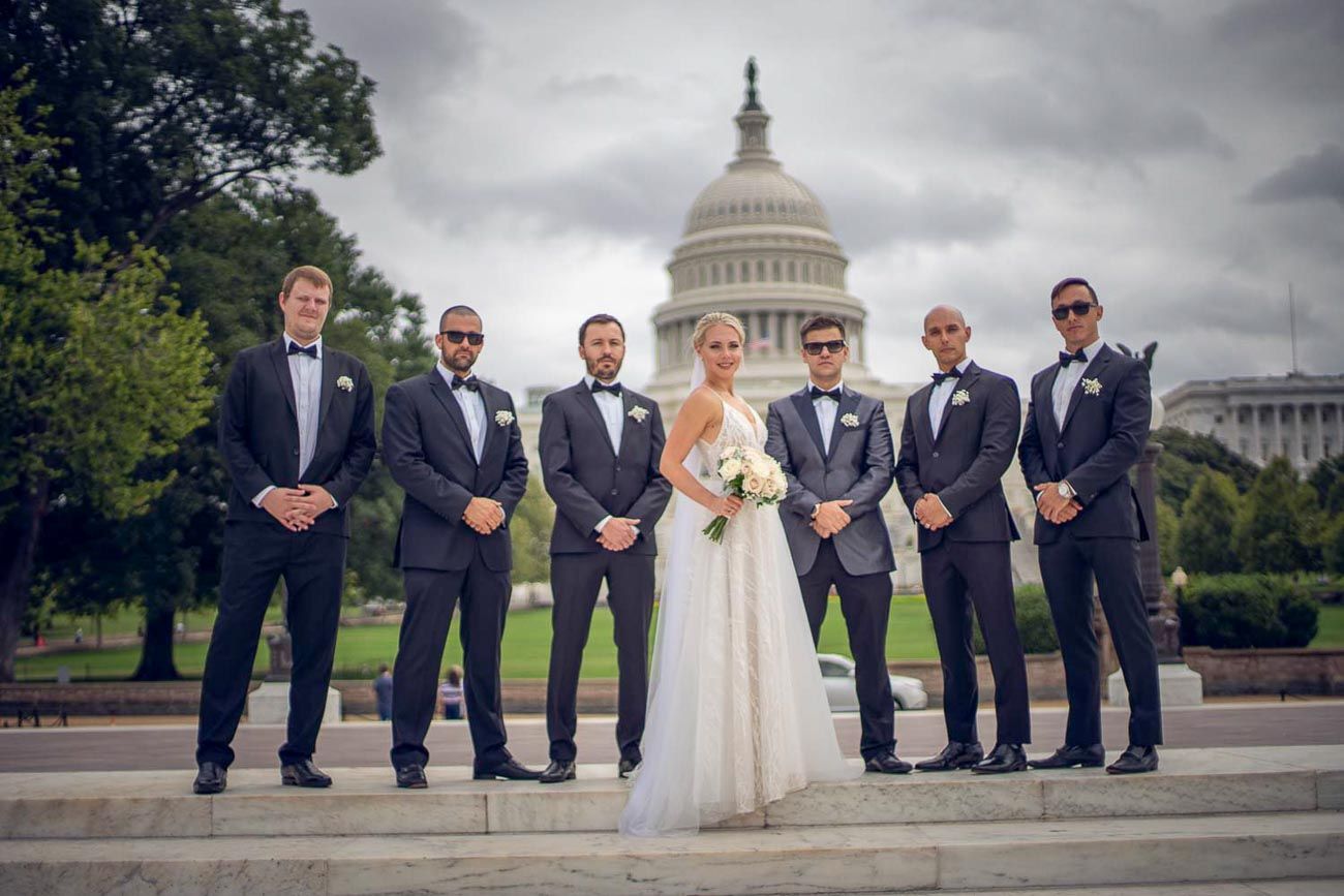 Wedding party poses on Capitol steps, bride in white gown with groomsmen in dark tuxedos.
