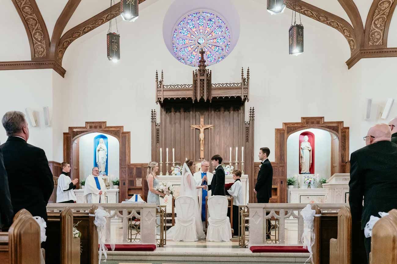 Bride and groom exchanging vows at the altar of a Catholic church with wedding party present.