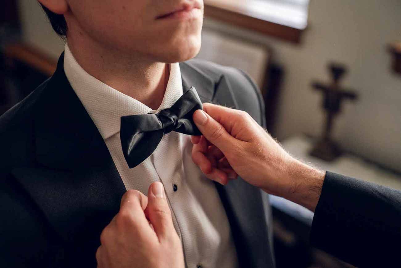 Close-up of hands adjusting a black bow tie on a groom in a formal tuxedo.