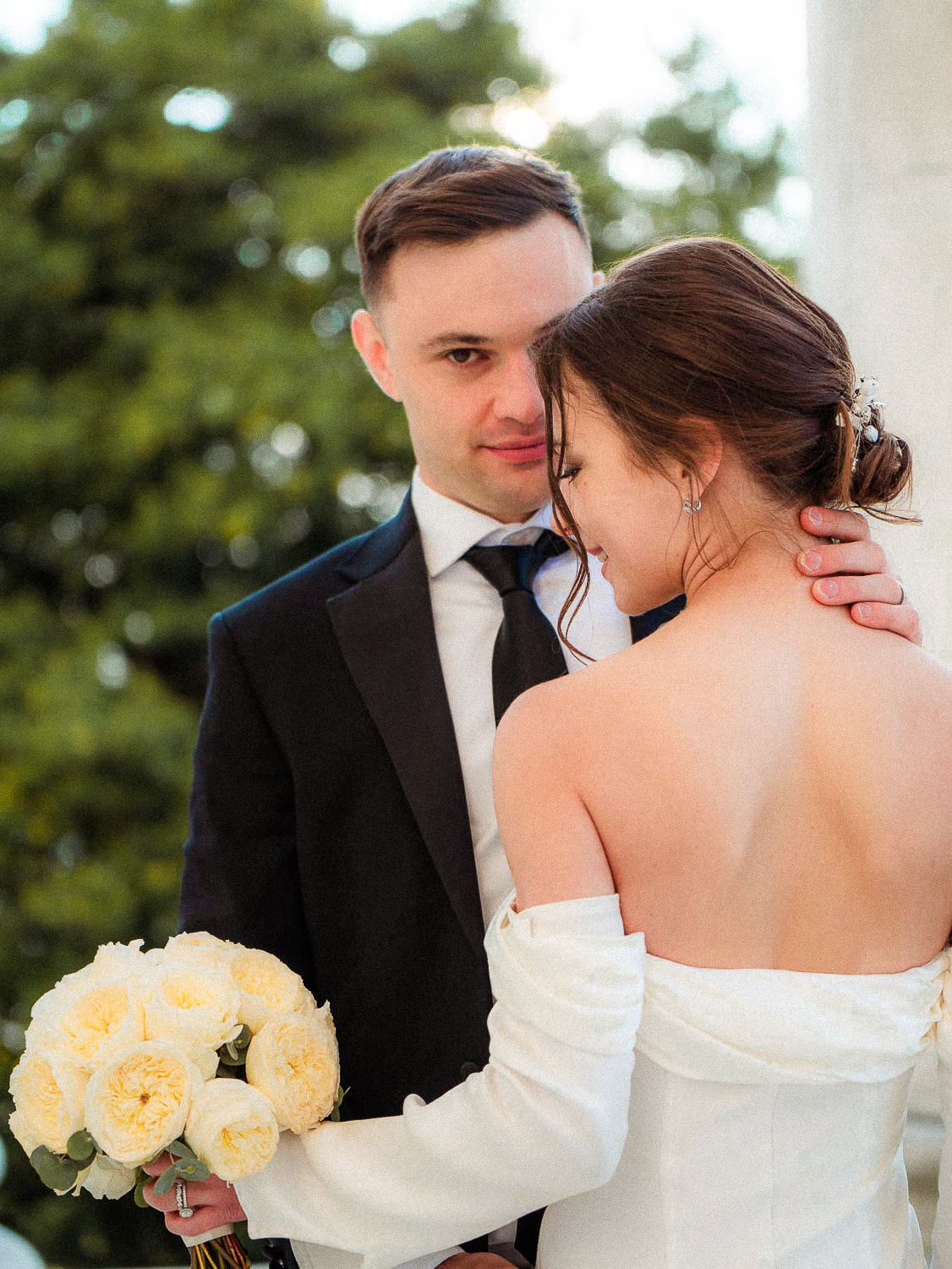 Groom in black tuxedo embraces bride in off-shoulder gown while holding white bouquet outdoors.