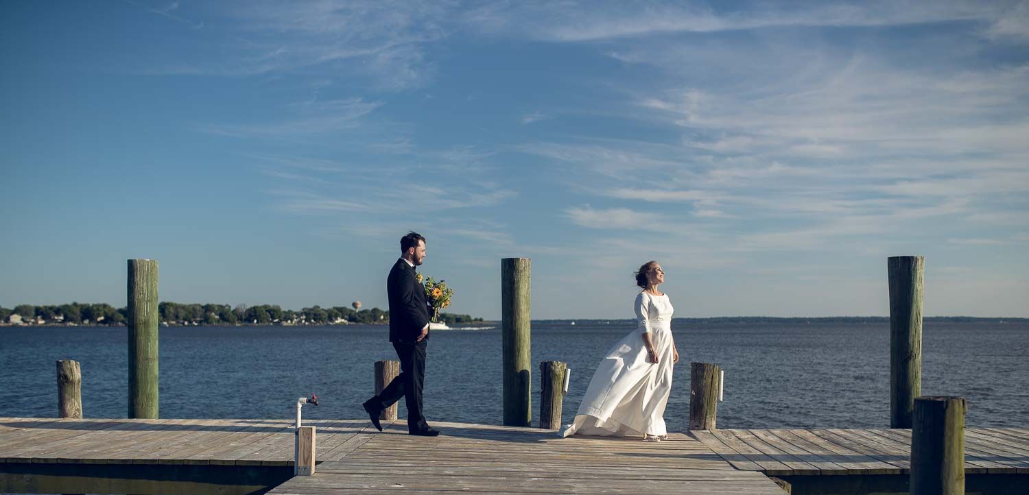 Bride and groom walking on a wooden dock by the water under a bright blue sky on their wedding day.