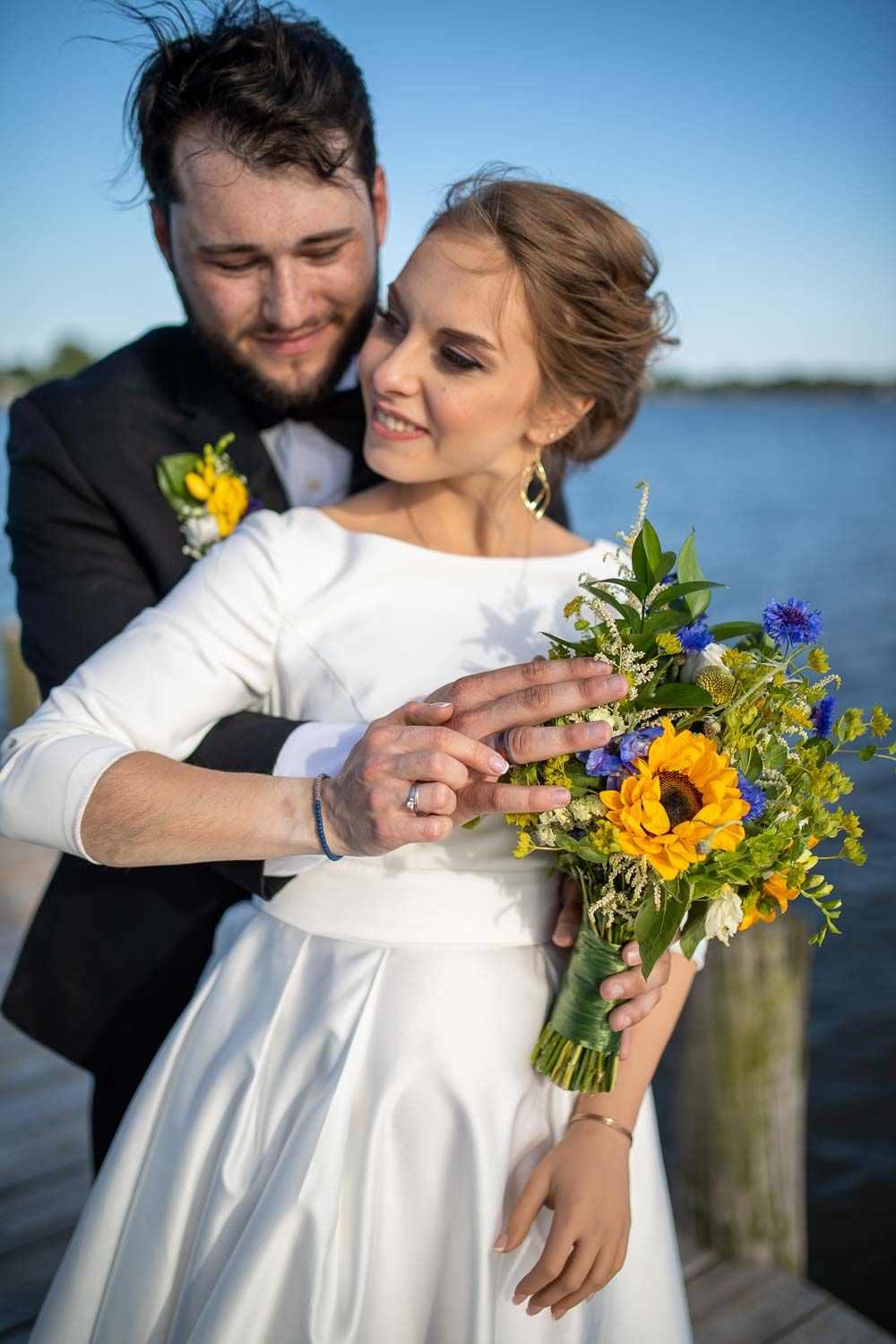 Smiling bride in white gown held by groom in tuxedo, showcasing wedding rings and sunflower bouquet.