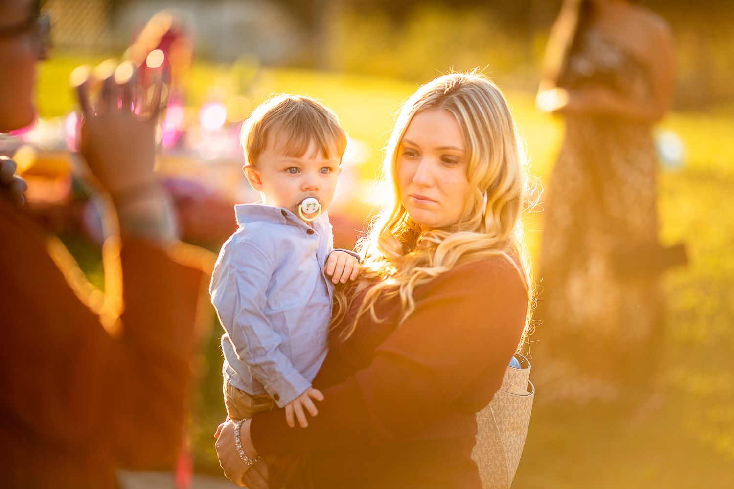 Blonde woman holding a baby with pacifier during an outdoor golden hour celebration.