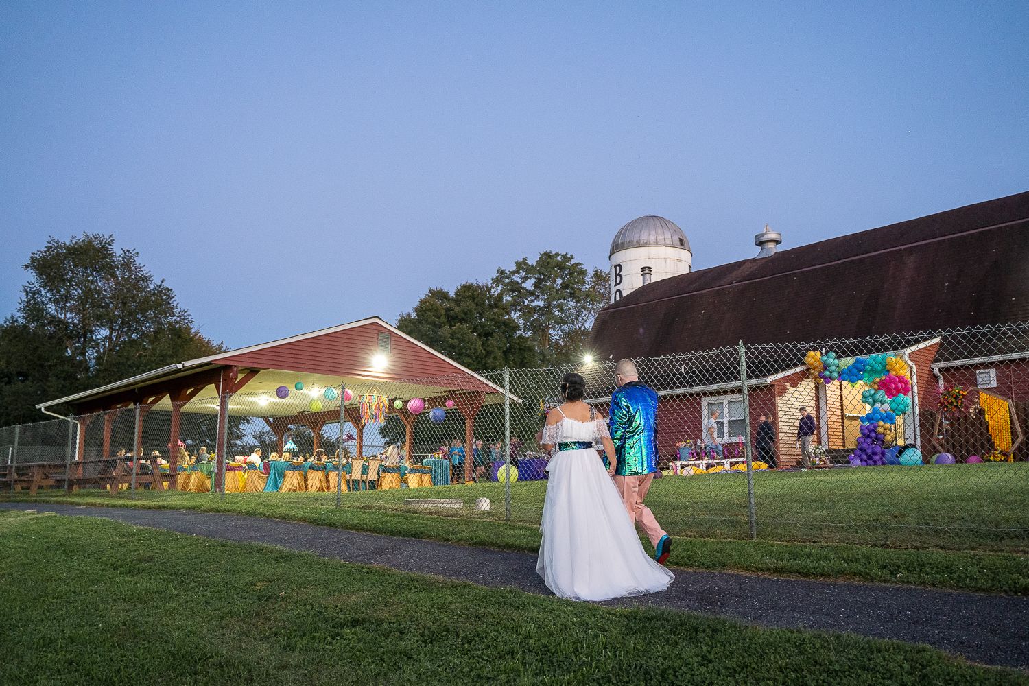 Bride in white gown stands before illuminated barn venue at dusk during outdoor wedding celebration.