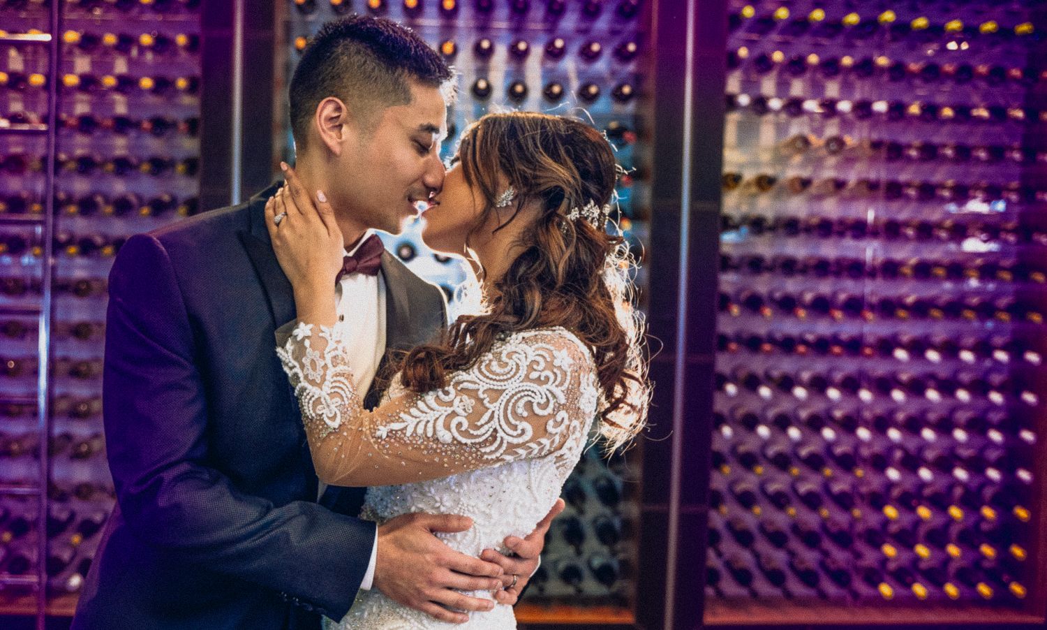 Bride and groom sharing a romantic kiss in front of a wine bottle display wall.