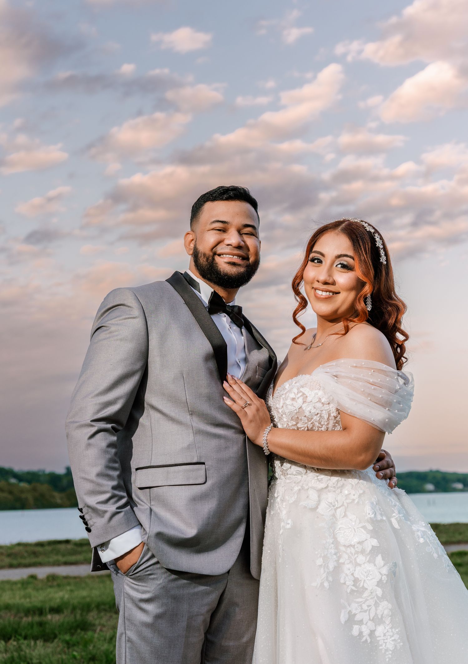 Couple posing outdoors at sunset in wedding attire, groom in gray tuxedo and bride in white lace off-shoulder gown.