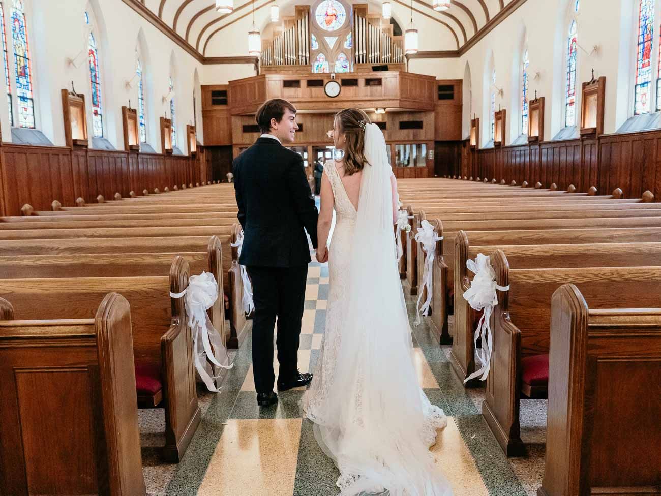 Newlyweds hold hands walking down the aisle of a classic church with white bow-decorated pews.