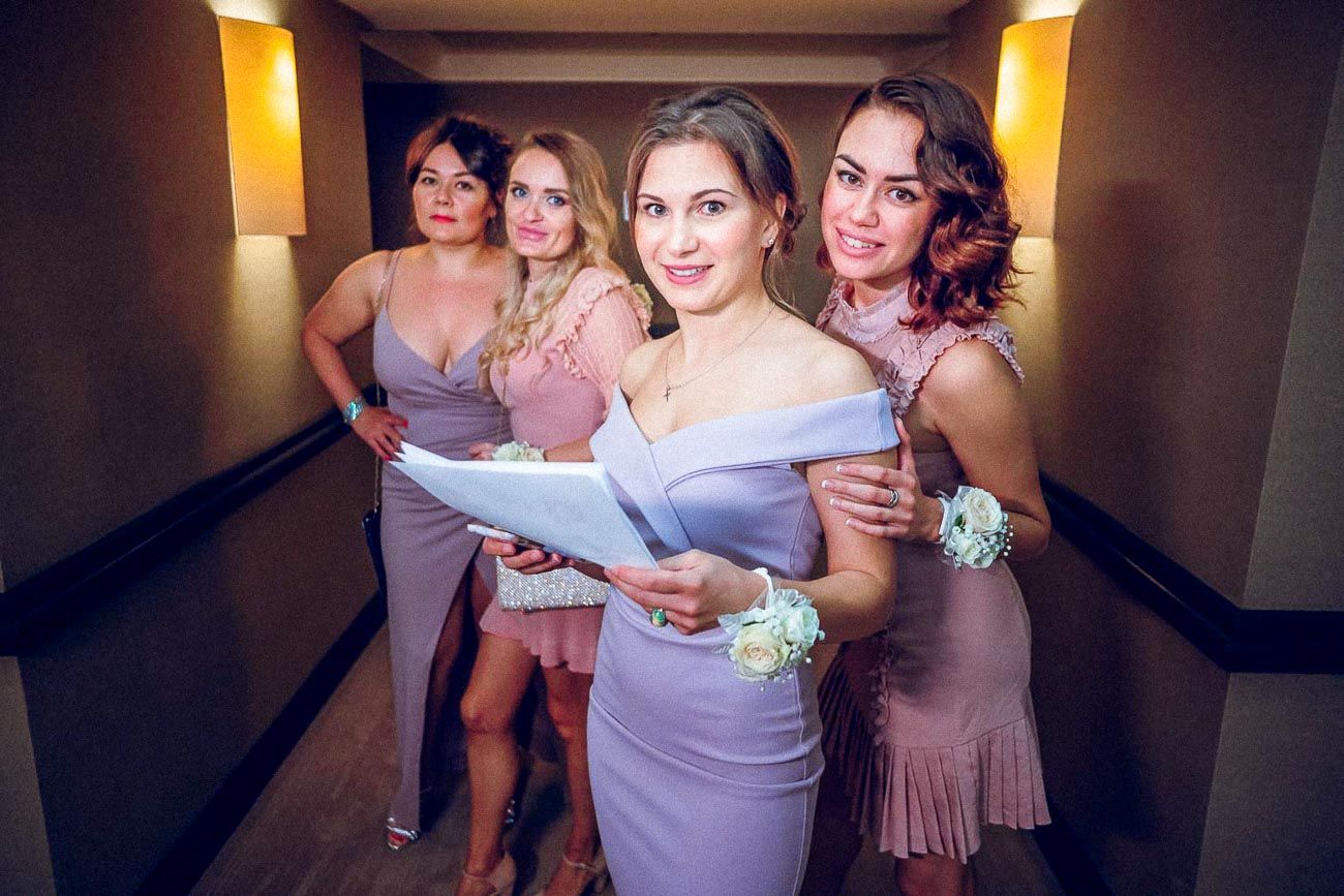 Four bridesmaids in lavender dresses posing together in hotel hallway holding flowers and papers.