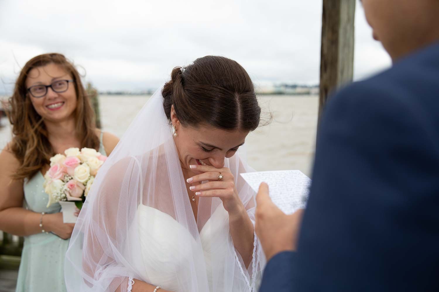 Emotional bride covers mouth while crying during wedding ceremony as bridesmaid watches nearby.