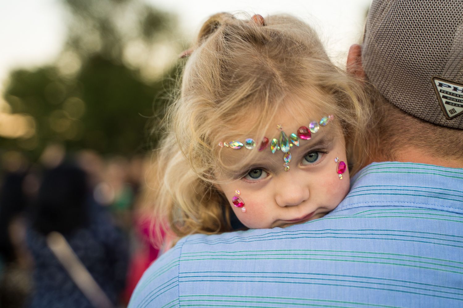 Young girl with glitter face paint rests head on adult's shoulder at an outdoor celebration.