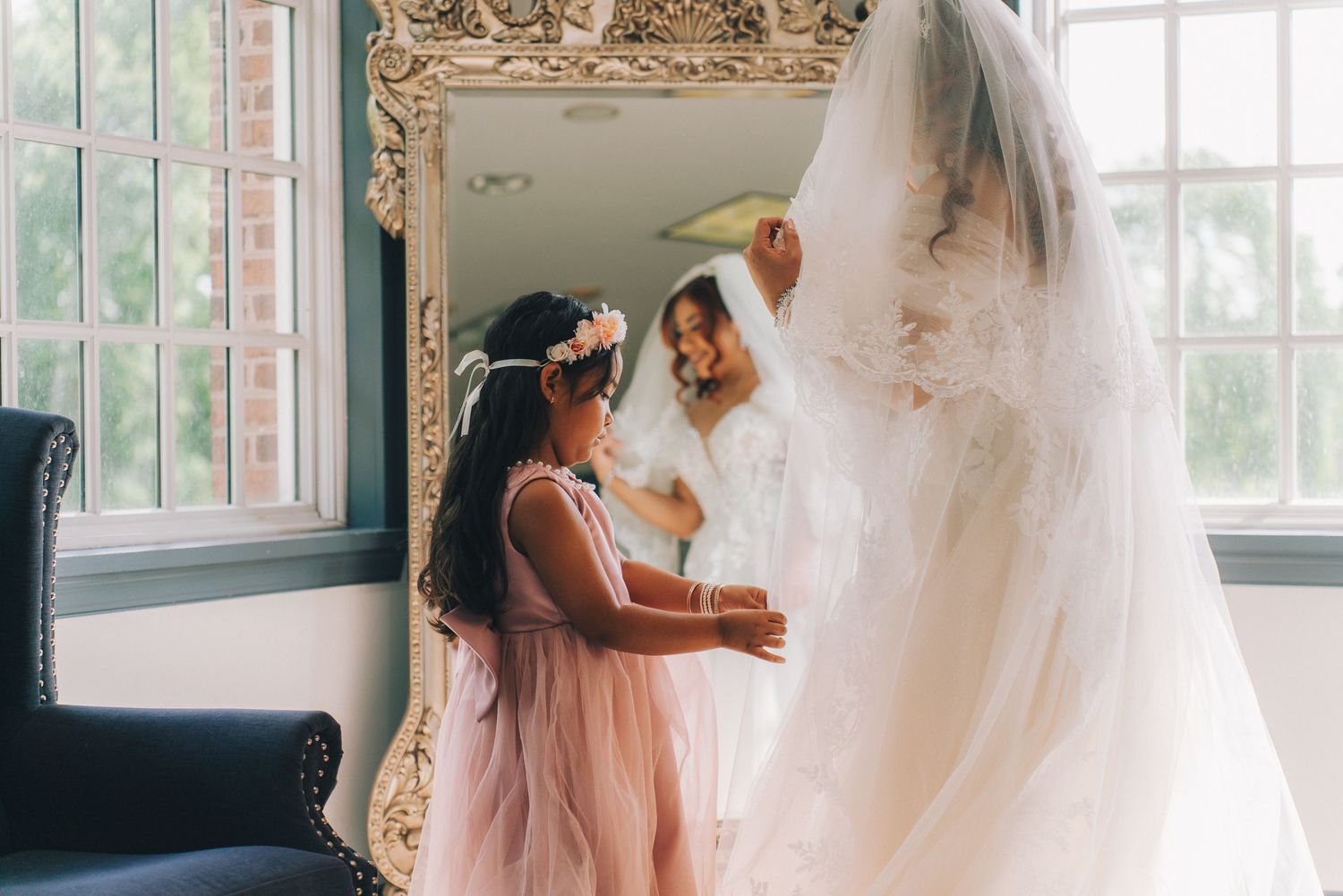 Flower girl in pink dress helps bride with wedding veil near ornate mirror in bright elegant bridal suite.