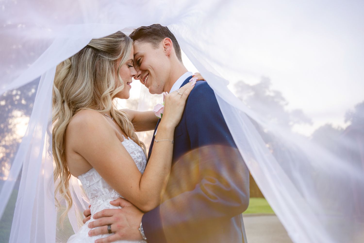 Bride and groom smiling under a flowing white veil on their wedding day.