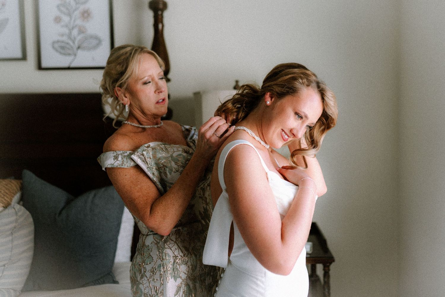 A mother helps her daughter into a white wedding dress in a softly lit bedroom before the ceremony.