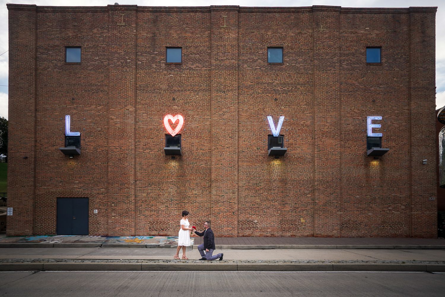 Man proposing on one knee to woman in front of a brick building with large LOVE neon sign letters on the wall.